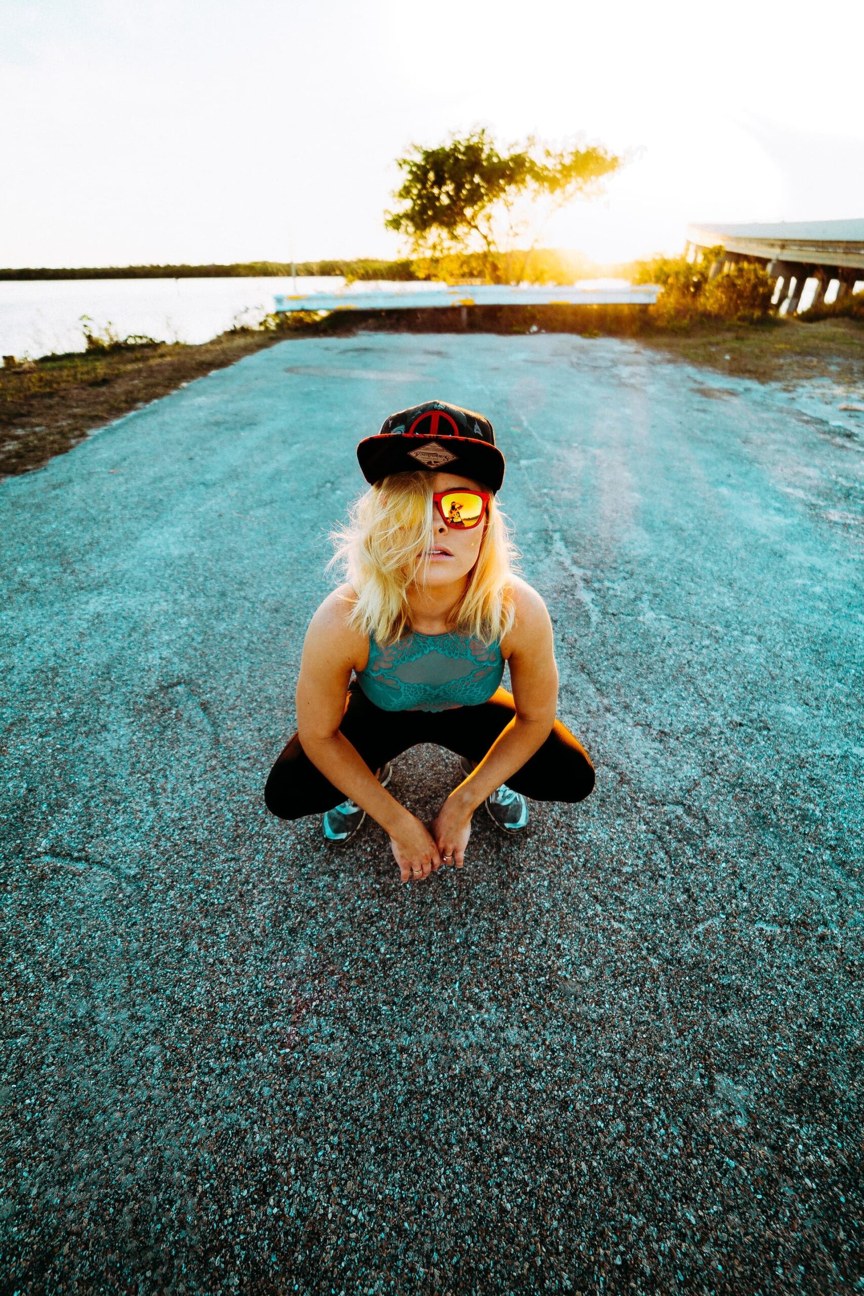 Beautiful blonde woman wearing a baseball cap and sunglasses crouching with a sunset in the back