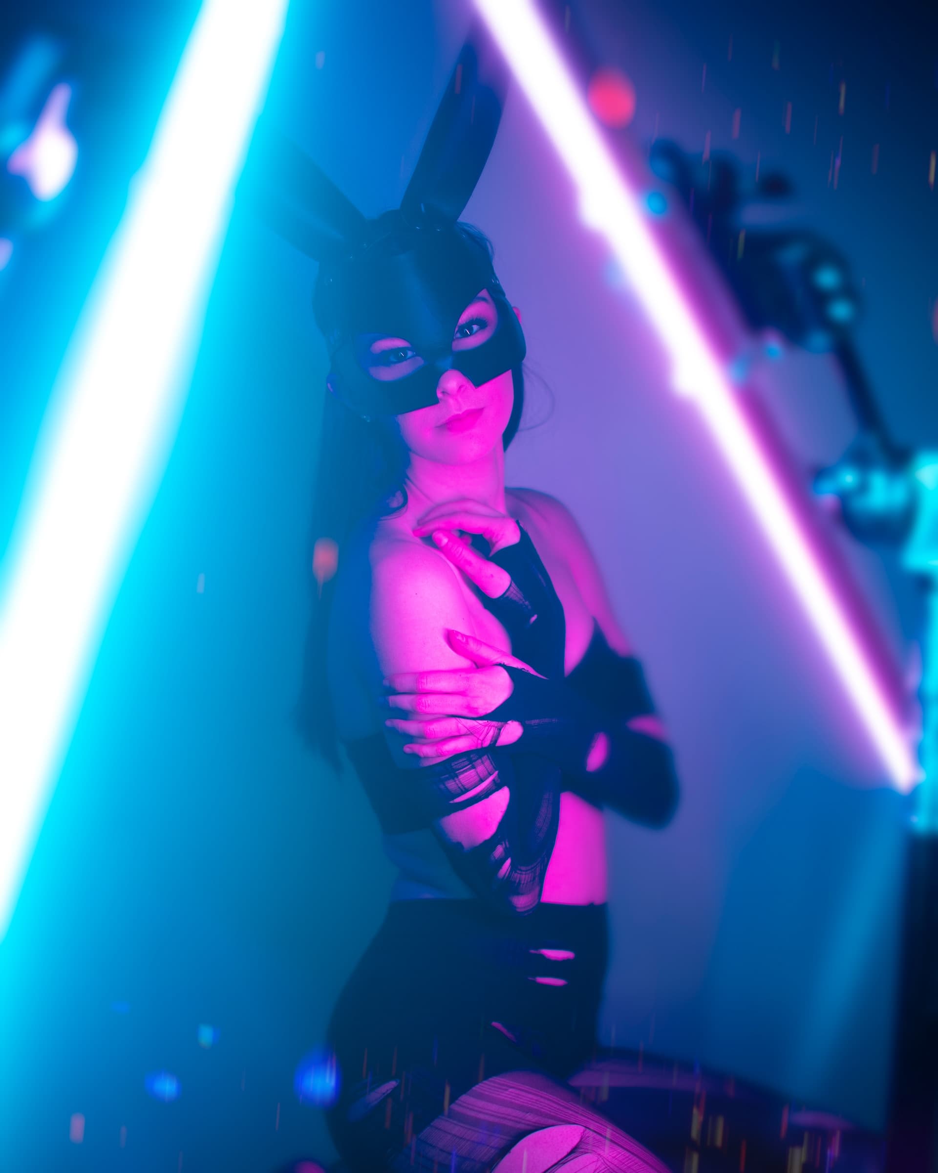 a woman wearing leather beds mask with bunny ears hiding her breast with her hands in a colourful neon lighting