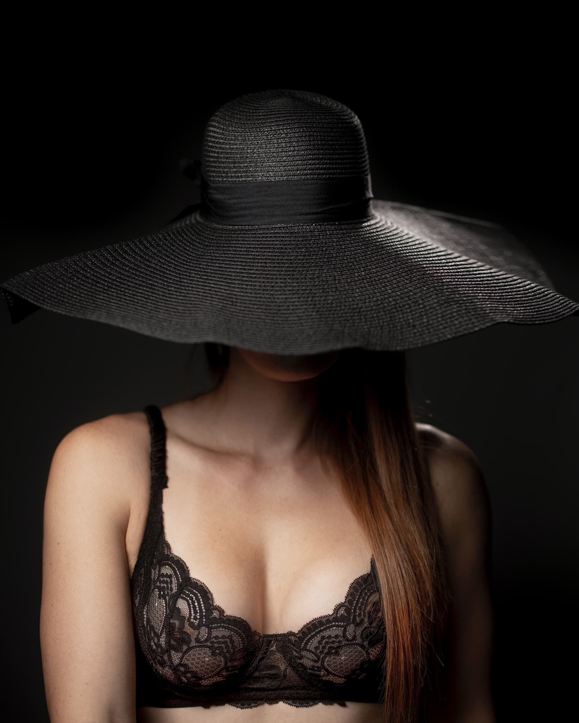 a woman wearing a black lace bra showing her cleavage is hiding her face with a large black hat