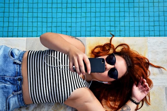 woman lying by the pool wearing sunglasses holding a phone on a sunny day