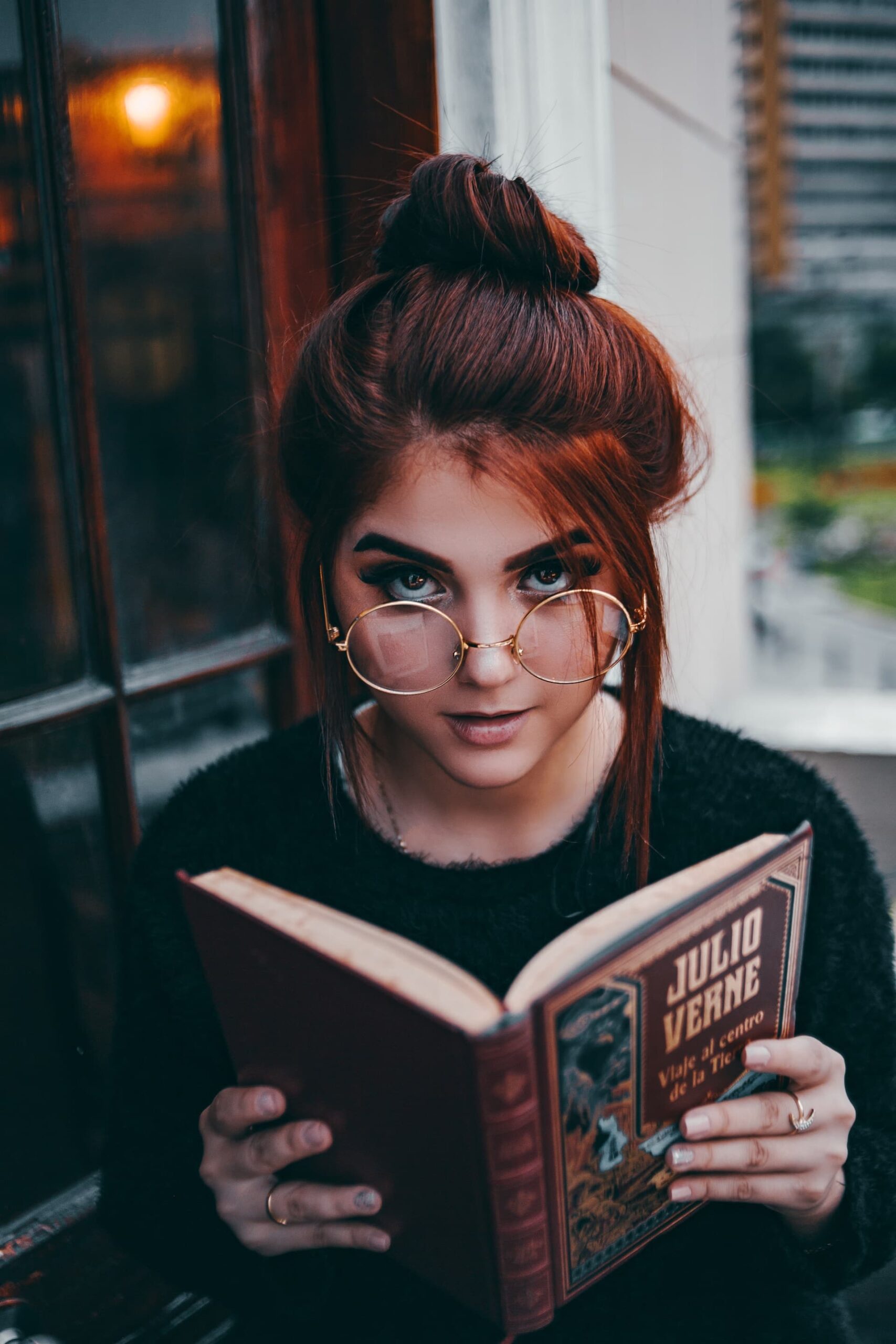 beautiful red hair woman with big round glasses reading a Jules Verne book
