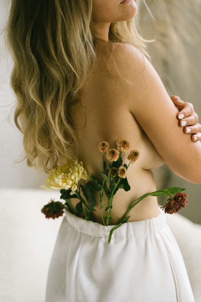 implied nude of a blonde model shot from the back. she has her arms crossed with flowers in her white pants