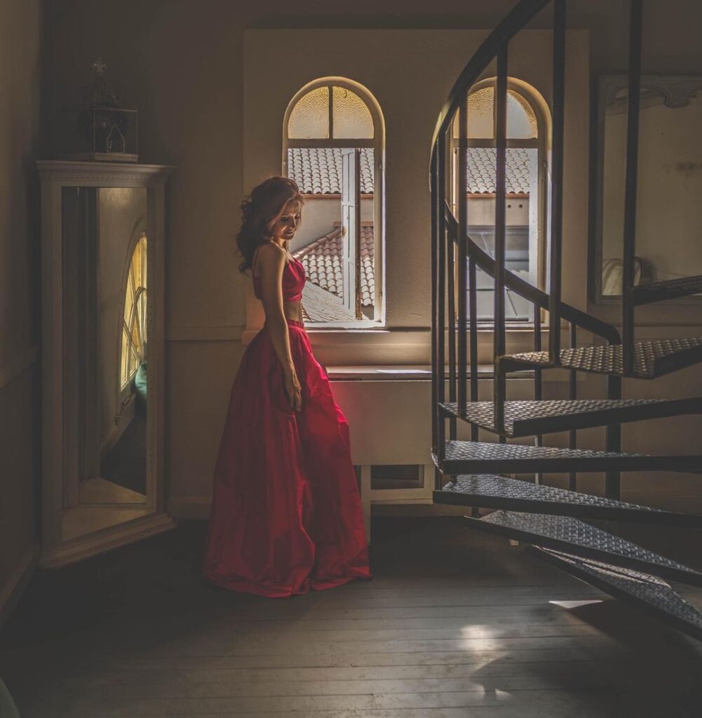 Woman wearing a classy long red dress standing in a majestic hall with a spinning staircase. Photo by Rita Hough