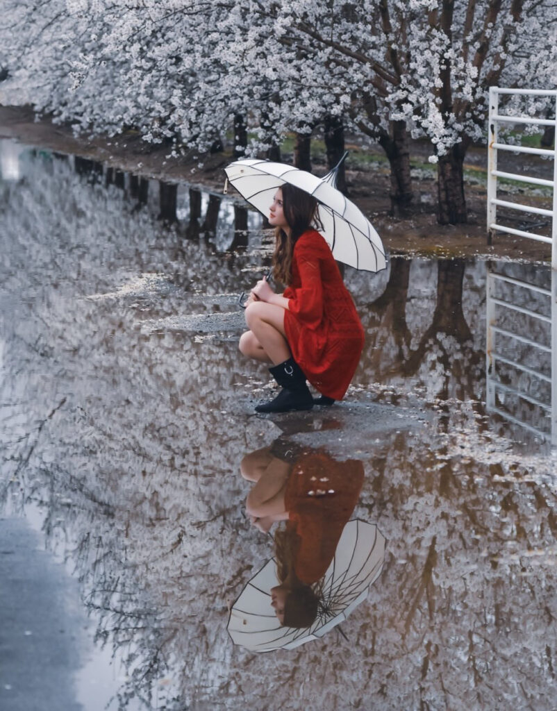 beautiful woman wearing a red rain coat and black boots crouching holding an umbrella. Photo by Rita Hough