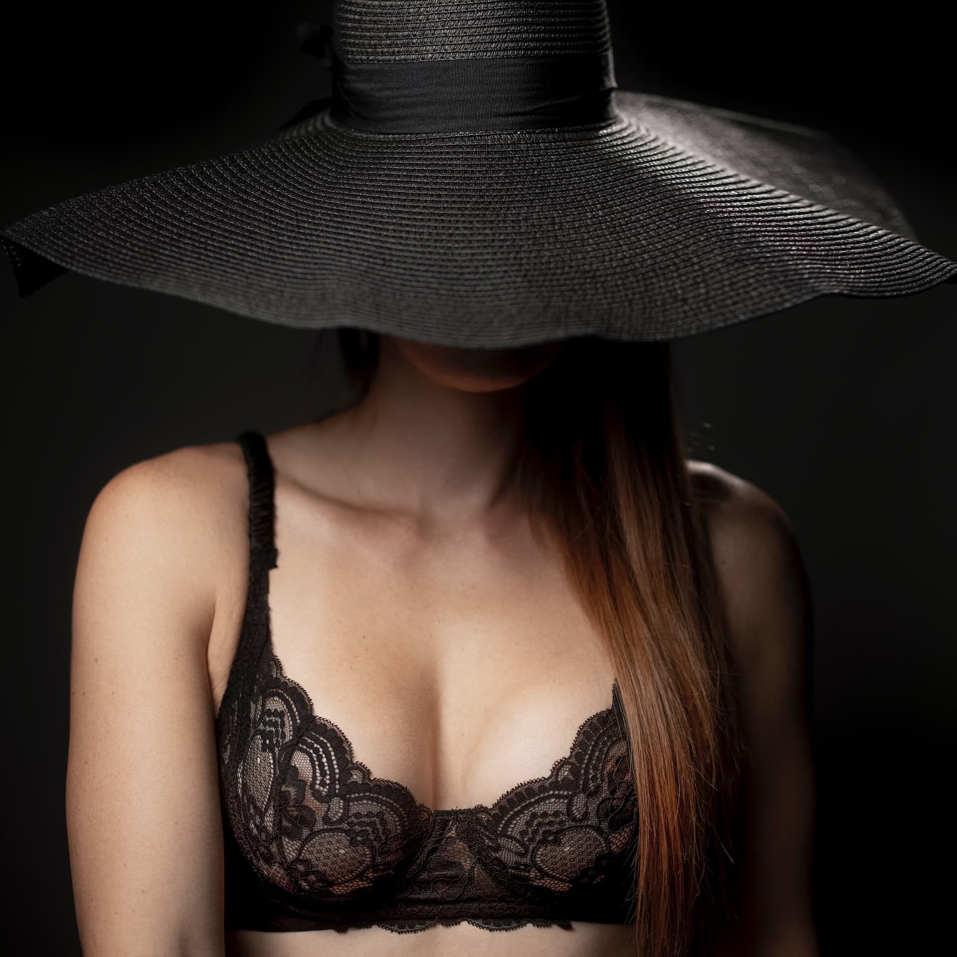 a woman wearing a black lace bra showing her cleavage is hiding her face with a large black hat